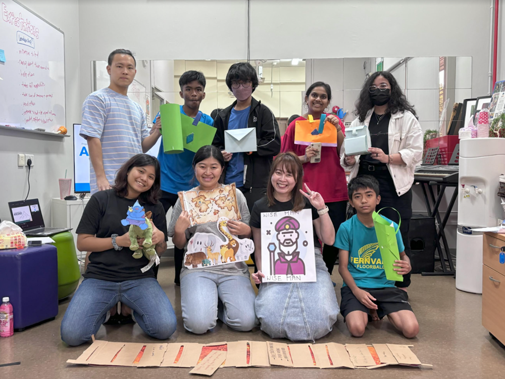 Group of nine people holding crafts like paper bags and drawings in an indoor space.